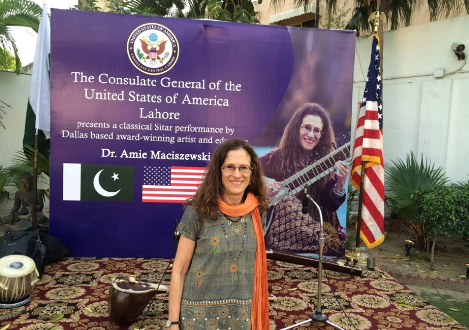 Amie in front of the official poster announcing her sitar concert at the residence of the U.S. Consulate General in Lahore. The poster bears the seal of the U.S.